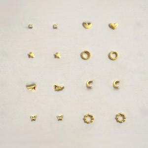 Pode incluir: Uma coleção de dezasseis brincos de pino dourados dispostos em quatro filas. O conjunto inclui pares de pequenos brincos de cristal, formas de coração, estrelas, círculos texturizados, asas, luas crescentes, borboletas e aros com missangas.