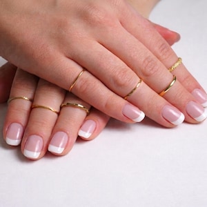 Op de afbeelding: Close-up van handen met French manicure nagels en meerdere gouden ringen. De ringen hebben verschillende ontwerpen, waaronder gladde banden en getextureerde patronen. De nagels hebben een roze basis met witte uiteinden.