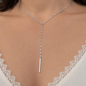 May include: A silver Y-shaped necklace with a delicate chain and a vertical bar pendant. The necklace is worn against a white top with lace trim, creating a simple and elegant look. The necklace is a stylish accessory.