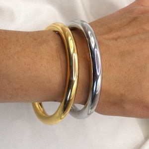 Op de afbeelding: Twee metalen armbanden, één goudkleurig en één zilverkleurig, worden om een pols gedragen. De gouden armband heeft een gepolijste afwerking, terwijl de zilveren armband licht reflecteert. De sieraden worden tegen een witte achtergrond getoond.