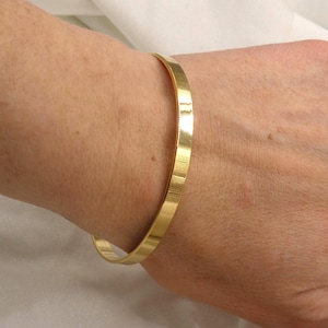 Può includere: Un bracciale rigido in metallo color oro con una finitura liscia e lucida. Il bracciale presenta un design a fascia sottile e piatta che circonda il polso, mostrando una superficie riflettente che cattura la luce lungo tutta la circonferenza.