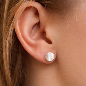 Peut inclure: Gros plan d'une boucle d'oreille argentée en forme de cercle. La boucle d'oreille est dans l'oreille d'une personne aux cheveux blonds. La boucle d'oreille a une finition en métal brossé.