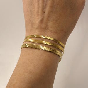 Peut inclure: Trois bracelets en chaîne serpent dorés superposés au poignet d'une personne. Les bracelets sont en métal brillant et lisse.