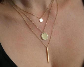 Puede incluir: Un conjunto de collares dorados en capas que presenta tres cadenas delicadas de diferentes longitudes. La cadena más corta sostiene un pequeño disco circular, la cadena intermedia presenta un disco circular más grande y la cadena más larga muestra un colgante de barra vertical.
