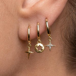 Peut inclure: Gros plan sur trois boucles d'oreilles dorées. Chaque boucle d'oreille a un charme différent : une étoile à quatre branches, une découpe en forme d'étoile et une étoile à quatre branches avec de petites pierres claires. Les boucles d'oreilles sont portées sur une oreille.