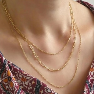 Könnte beinhalten: Ein mehrschichtiges goldenes Halsketten-Set. Die Halsketten variieren im Stil, mit zarten Ketten und größeren Gliederketten-Designs. Der Schmuck wird vor einem gemusterten bordeauxroten und cremefarbenen Stoff präsentiert.