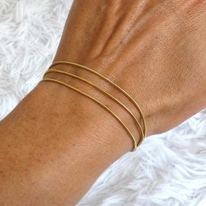 Op de afbeelding: Een set van drie dunne, goudkleurige metalen armbanden met een glad, afgerond oppervlak. De armbanden worden samen gedragen, wat zorgt voor een gestapelde uitstraling om de pols, met een eenvoudig, doorlopend cirkelvormig ontwerp.