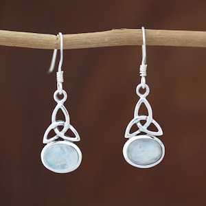 May include: Silver Celtic knot earrings with a white moonstone cabochon. The earrings are hanging from a branch.