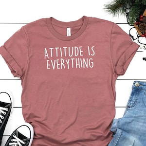 May include: A mauve t-shirt with the text "ATTITUDE IS EVERYTHING" in white lettering. The t-shirt is laid flat on a white wooden surface with a pair of black Converse sneakers and blue jeans with distressed details.