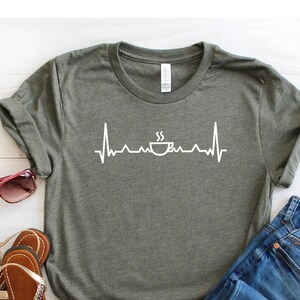 Puede incluir: Una camiseta verde con un gr&aacute;fico blanco de una taza de caf&eacute; con vapor saliendo de ella, dentro de una l&iacute;nea de latido del coraz&oacute;n. La camiseta est&aacute; colocada sobre una superficie de madera blanca con un par de sandalias marrones y unos vaqueros azules.