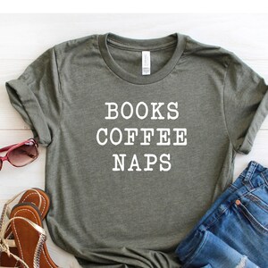 May include: A green t-shirt with the text "BOOKS COFFEE NAPS" in white lettering. The shirt is paired with blue jeans and brown sandals.