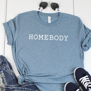 Pode incluir: Uma camiseta azul com a palavra "HOMEBODY" impressa em branco na frente. A camiseta est&aacute; colocada sobre uma superf&iacute;cie de madeira branca com um par de cal&ccedil;as de ganga azuis e um par de t&eacute;nis azul escuro.