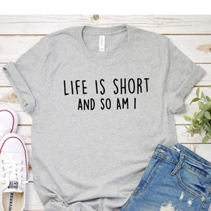 May include: A gray t-shirt with the text "Life is short and so am I" printed on it. The shirt is laid out on a white wooden surface with a pair of white sneakers and denim shorts.