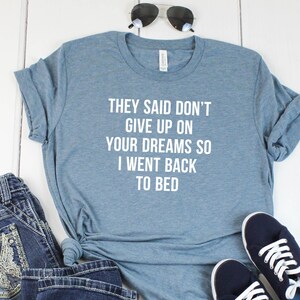 May include: A blue t-shirt with the text "They said don't give up on your dreams so I went back to bed" printed in white. The shirt is laid out on a white wooden surface with a pair of blue jeans, sunglasses, and a pair of blue sneakers.