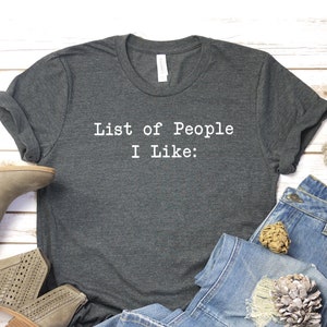 May include: A dark gray t-shirt with the text "List of People I Like:" printed in white on the front. The shirt is laid out on a white wooden surface with a pair of light brown suede ankle boots, a pair of blue jeans, and a decorative piece.