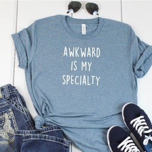 May include: A blue t-shirt with the text "Awkward is my specialty" printed in white on the front. The shirt is paired with blue jeans and dark blue sneakers.