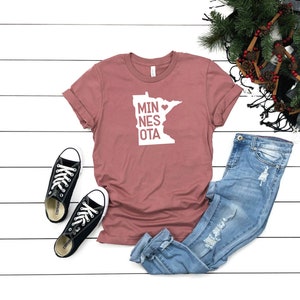 May include: A dusty rose t-shirt with a white graphic of the state of Minnesota with a heart in the middle and the text "MINNESOTA" written vertically. The t-shirt is laid out on a white wooden surface with a pair of black Converse sneakers, blue jeans, and a brown and green scarf with pine needles and cinnamon sticks.