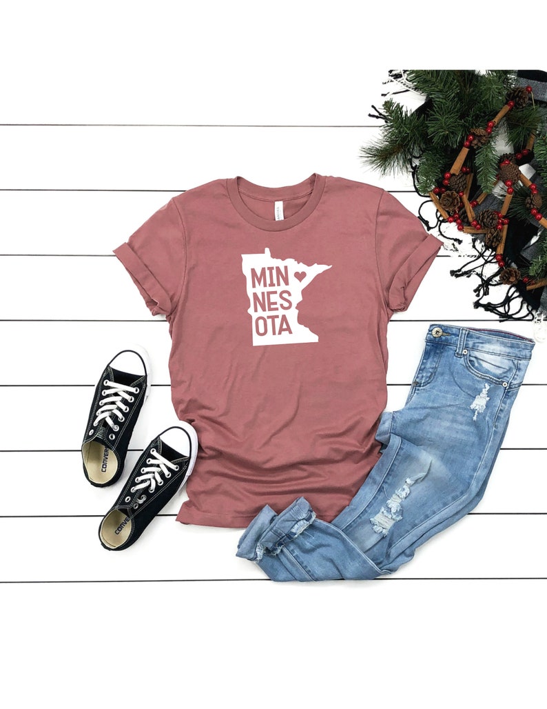 May include: A dusty rose t-shirt with a white graphic of the state of Minnesota with a heart in the middle and the text "MINNESOTA" written vertically. The t-shirt is laid out on a white wooden surface with a pair of black Converse sneakers, blue jeans, and a brown and green scarf with pine needles and cinnamon sticks.