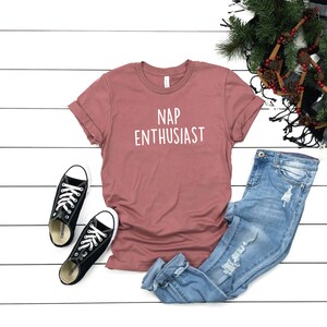 May include: A dusty rose t-shirt with the text "NAP ENTHUSIAST" in white. The t-shirt is laid flat on a white wooden surface with a pair of black Converse sneakers and blue jeans.