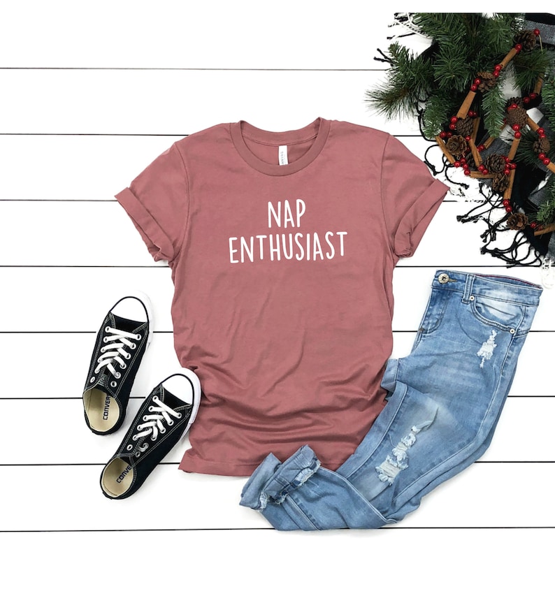 May include: A dusty rose t-shirt with the text "NAP ENTHUSIAST" in white. The t-shirt is laid flat on a white wooden surface with a pair of black Converse sneakers and blue jeans.
