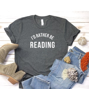 May include: A dark gray t-shirt with the text "I'd rather be reading" in white lettering. The shirt is laid out on a white wooden surface with a pair of light brown ankle boots, blue jeans, and decorative items.