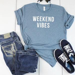May include: A blue t-shirt with the text "Weekend Vibes" in white lettering. The shirt is laid out on a white wooden surface with a pair of blue jeans, sunglasses, and a pair of blue sneakers.