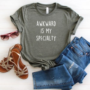 May include: A green t-shirt with the text "AWKWARD IS MY SPECIALTY" in white lettering. The shirt is paired with blue jeans and brown sandals.
