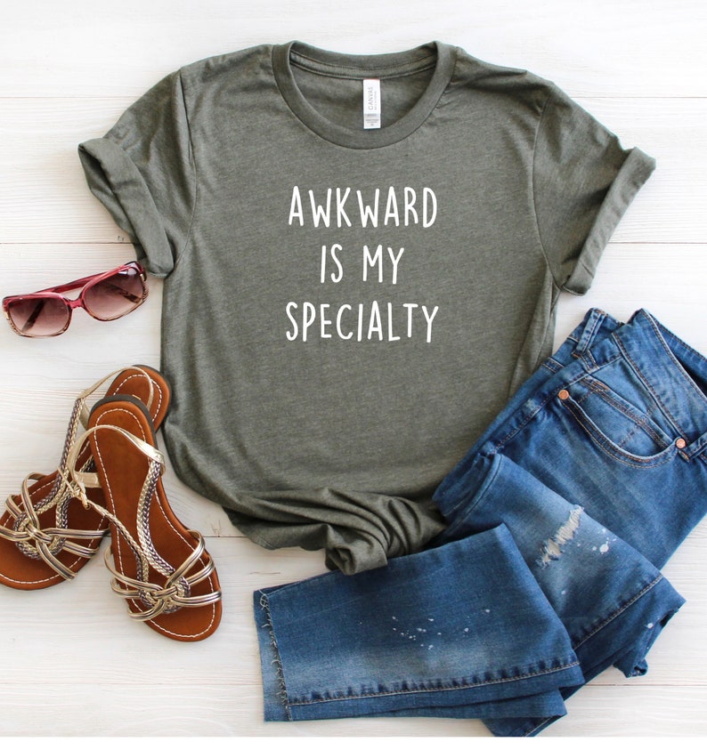 May include: A green t-shirt with the text "AWKWARD IS MY SPECIALTY" in white lettering. The shirt is paired with blue jeans and brown sandals.