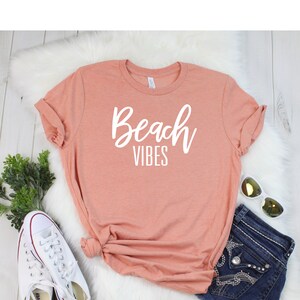 K&ouml;nnte beinhalten: Ein pfirsichfarbenes T-Shirt mit dem Text "Beach Vibes" in wei&szlig;en Buchstaben. Das Shirt liegt auf einer wei&szlig;en, flauschigen Oberfl&auml;che mit einem Paar wei&szlig;en Turnschuhen, einer blauen Jeans-Shorts und einer Sonnenbrille.