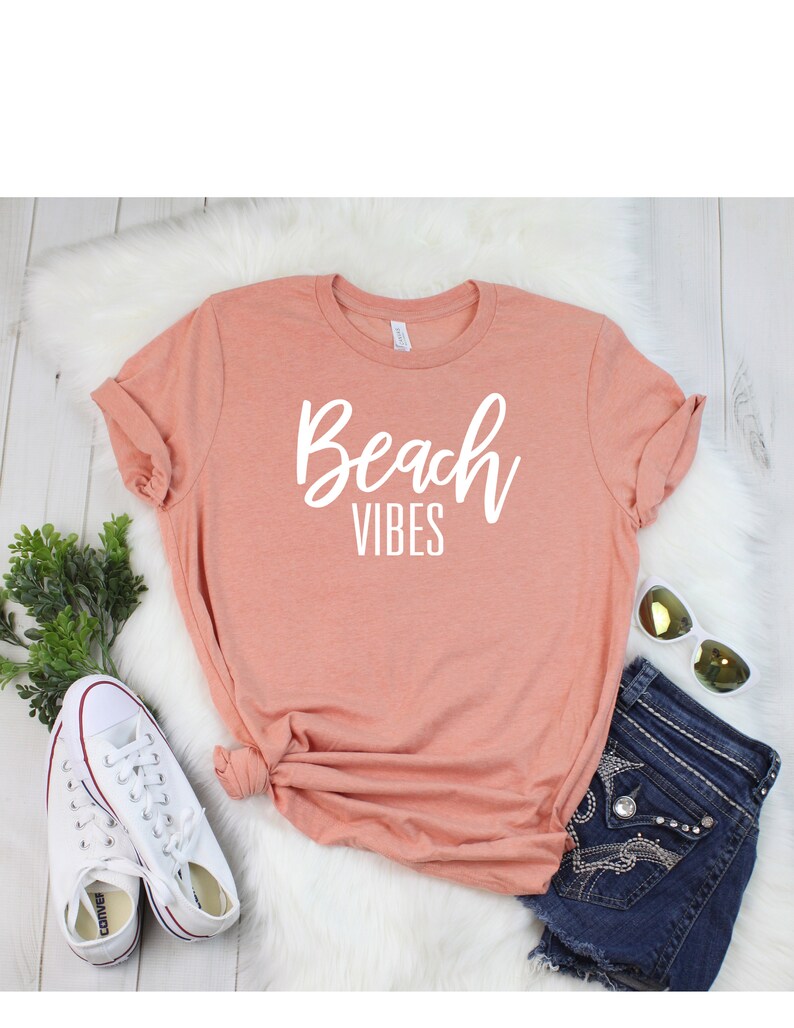 K&ouml;nnte beinhalten: Ein pfirsichfarbenes T-Shirt mit dem Text "Beach Vibes" in wei&szlig;en Buchstaben. Das Shirt liegt auf einer wei&szlig;en, flauschigen Oberfl&auml;che mit einem Paar wei&szlig;en Turnschuhen, einer blauen Jeans-Shorts und einer Sonnenbrille.