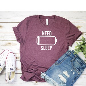 May include: A maroon t-shirt with the text "NEED SLEEP" and a white battery icon with a low battery level.