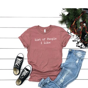 May include: A mauve t-shirt with the text "List of People I Like:" printed in white on the front. The shirt is paired with blue jeans and black sneakers.
