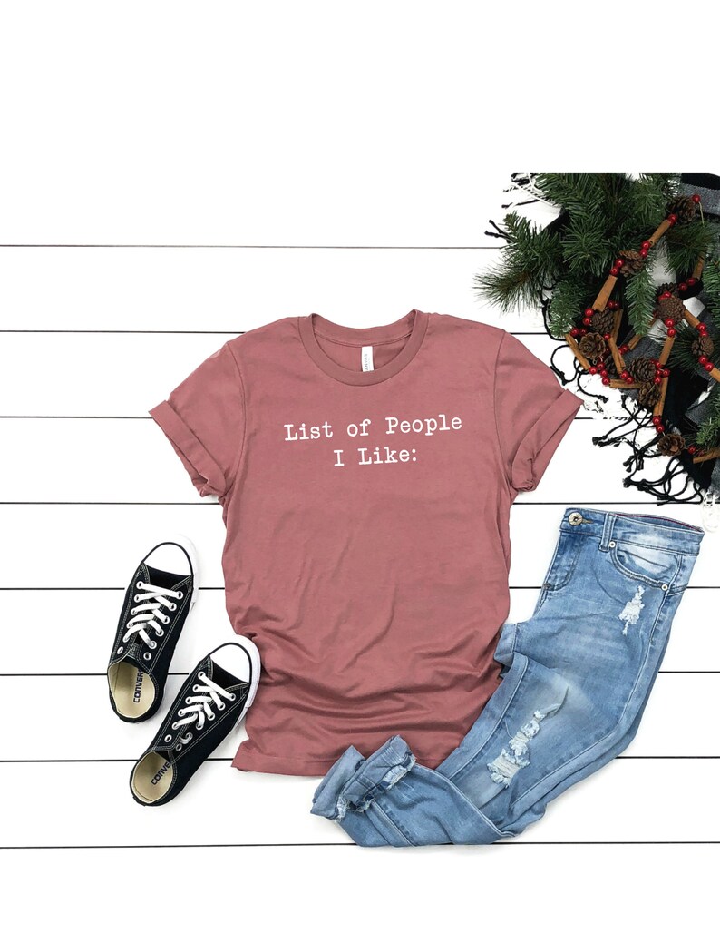 May include: A mauve t-shirt with the text "List of People I Like:" printed in white on the front. The shirt is paired with blue jeans and black sneakers.