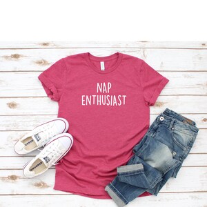May include: A pink t-shirt with the text "NAP ENTHUSIAST" in white lettering. The shirt is laid flat on a white wooden surface with a pair of white sneakers and blue jeans.