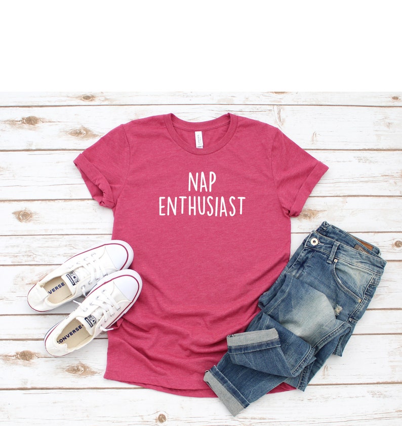 May include: A pink t-shirt with the text "NAP ENTHUSIAST" in white lettering. The shirt is laid flat on a white wooden surface with a pair of white sneakers and blue jeans.