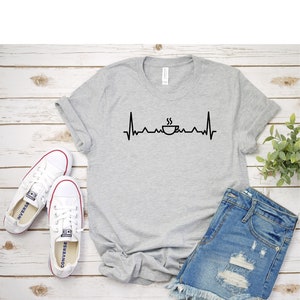 Puede incluir: Una camiseta gris con un gr&aacute;fico de taza de caf&eacute; negro y una l&iacute;nea de latido del coraz&oacute;n. La camiseta est&aacute; colocada sobre una superficie de madera blanca con un par de zapatillas blancas y pantalones cortos de mezclilla azul.