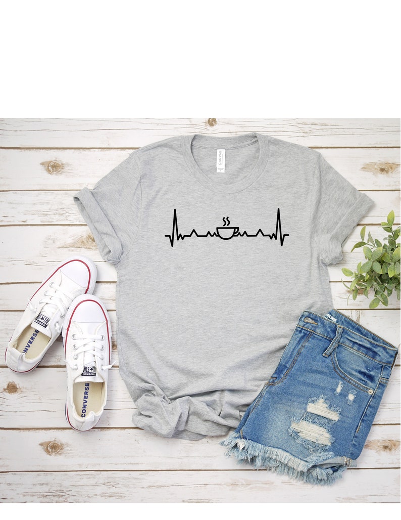 Puede incluir: Una camiseta gris con un gr&aacute;fico de taza de caf&eacute; negro y una l&iacute;nea de latido del coraz&oacute;n. La camiseta est&aacute; colocada sobre una superficie de madera blanca con un par de zapatillas blancas y pantalones cortos de mezclilla azul.