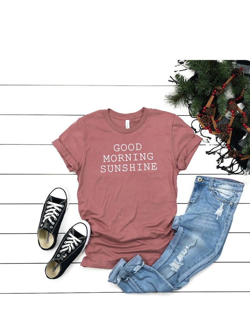 May include: A mauve t-shirt with the text "GOOD MORNING SUNSHINE" in white. The t-shirt is paired with blue jeans and black sneakers.