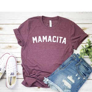 Pu&ograve; includere: Una maglietta color granata con la scritta "MAMACITA" stampata in bianco sul davanti. La maglietta &egrave; appoggiata su una superficie di legno bianco con un paio di scarpe da ginnastica bianche e un paio di pantaloncini di jeans blu.