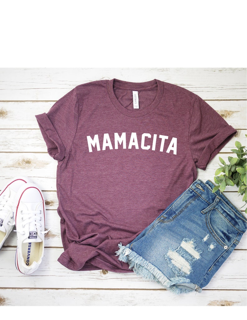 Pu&ograve; includere: Una maglietta color granata con la scritta "MAMACITA" stampata in bianco sul davanti. La maglietta &egrave; appoggiata su una superficie di legno bianco con un paio di scarpe da ginnastica bianche e un paio di pantaloncini di jeans blu.