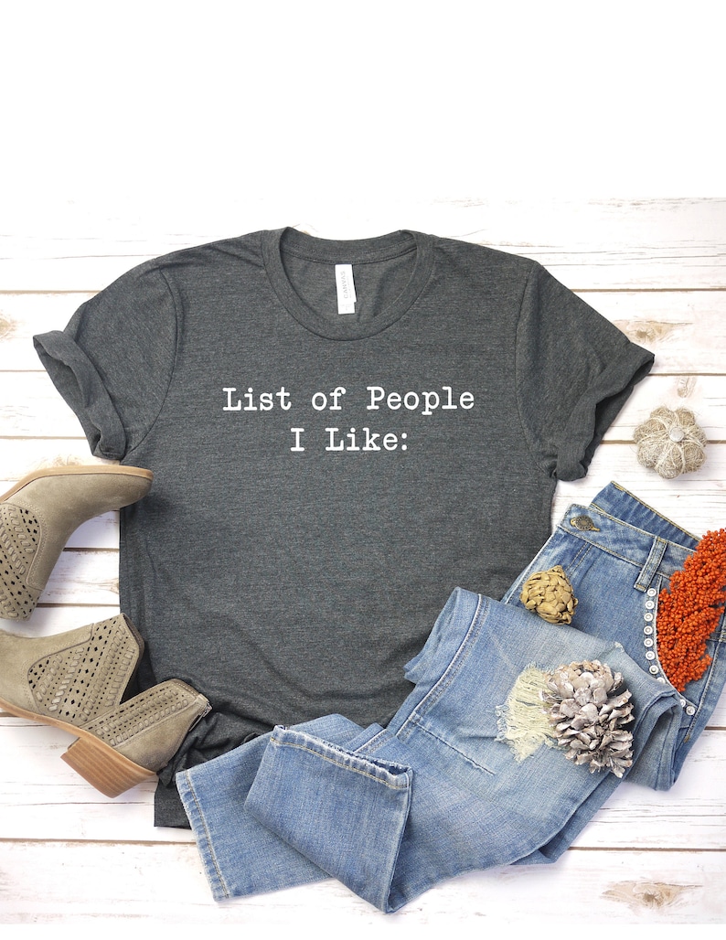 May include: A dark gray t-shirt with the text "List of People I Like:" printed in white on the front. The shirt is laid out on a white wooden surface with a pair of light brown suede ankle boots, a pair of blue jeans, and a decorative piece.
