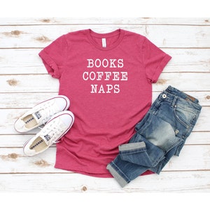 May include: A pink t-shirt with the text "BOOKS COFFEE NAPS" in white lettering. The shirt is laid out on a white wooden surface with a pair of white sneakers and blue jeans.