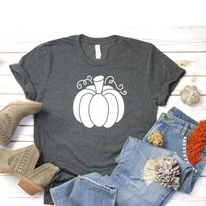 May include: A dark gray t-shirt with a white outline of a pumpkin with a stem and curly vines. The shirt is laid out on a white wooden surface with a pair of light brown ankle boots and blue jeans.
