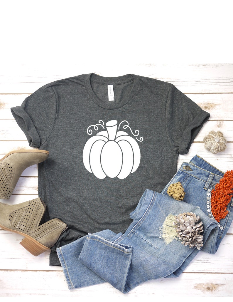 May include: A dark gray t-shirt with a white outline of a pumpkin with a stem and curly vines. The shirt is laid out on a white wooden surface with a pair of light brown ankle boots and blue jeans.