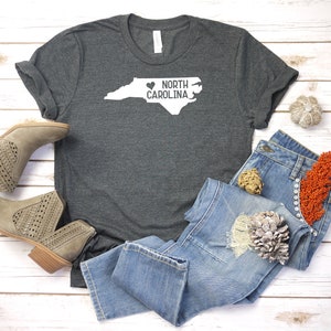 May include: A dark gray t-shirt with a white outline of the state of North Carolina and the text "NORTH CAROLINA" with a heart symbol in front of the word "NORTH".