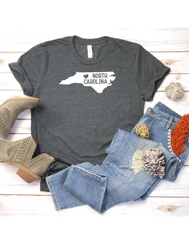 May include: A dark gray t-shirt with a white outline of the state of North Carolina and the text "NORTH CAROLINA" with a heart symbol in front of the word "NORTH".