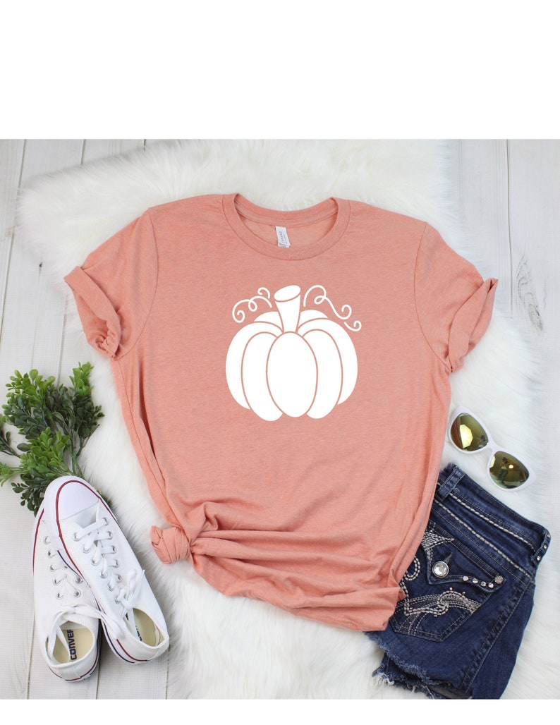 May include: A peach colored t-shirt with a white pumpkin graphic. The pumpkin has a stem and curly vines.