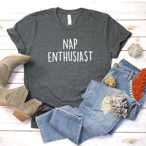 May include: A dark gray t-shirt with the text "NAP ENTHUSIAST" printed in white. The shirt is laid flat on a white wooden surface with a pair of brown ankle boots and blue jeans.