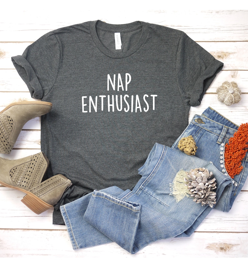 May include: A dark gray t-shirt with the text "NAP ENTHUSIAST" printed in white. The shirt is laid flat on a white wooden surface with a pair of brown ankle boots and blue jeans.