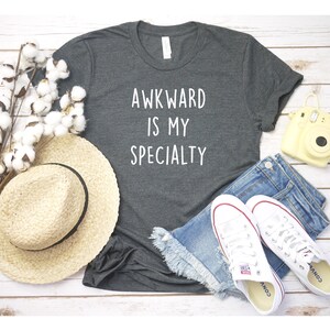 May include: A dark gray t-shirt with the text "AWKWARD IS MY SPECIALTY" in white lettering. The shirt is laid out on a white wooden surface with a straw hat, blue denim shorts, white sneakers, and a yellow camera.